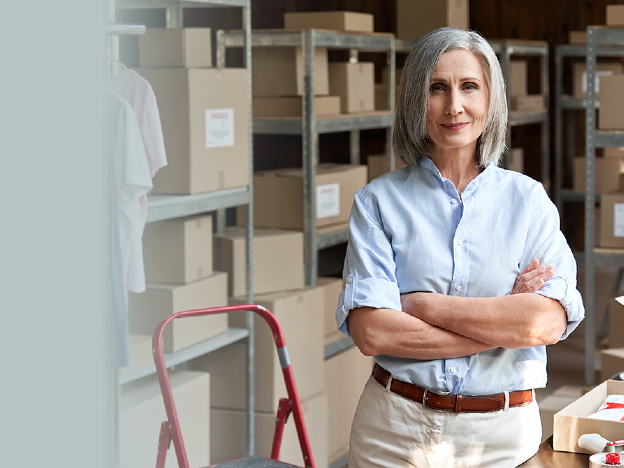 Confident happy mature older 60s woman retail seller, entrepreneur, clothing store small business owner, supervisor looking at camera standing arms crossed in delivery shipping warehouse, portrait.