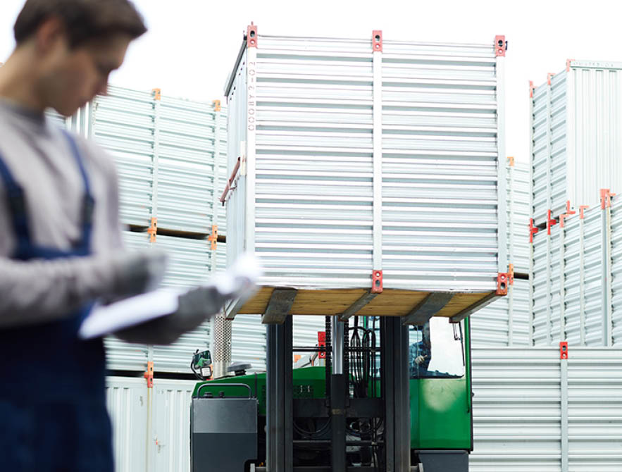 Powerful truck with heavy container moving along storage area between large containers, man controlling this process